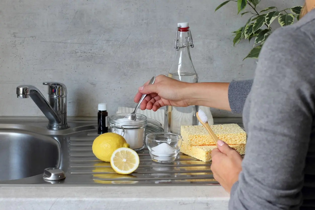 A view of someone making a DIY cleaner with baking soda, vinegar, a lemon, sponges, and a brush at the sink.