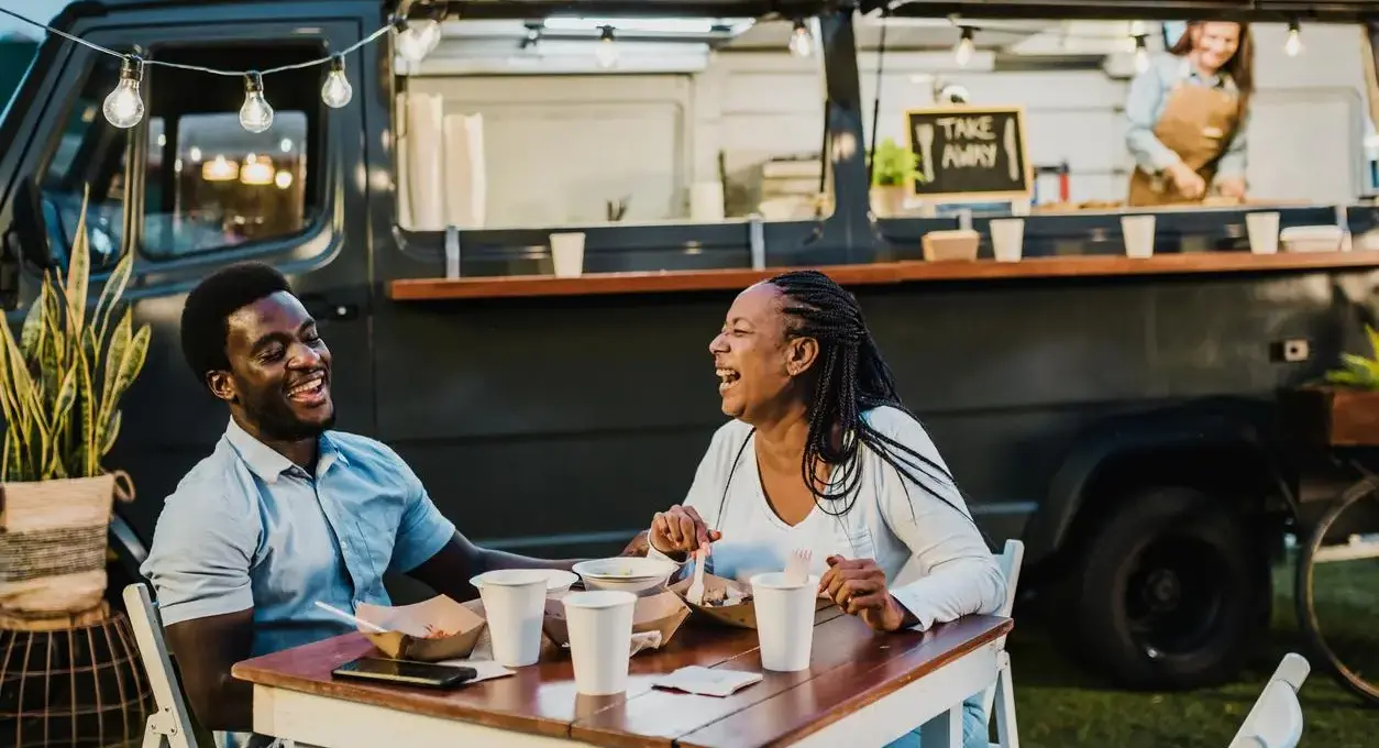 A laughing young couple enjoys a meal at a table in front of a food truck.