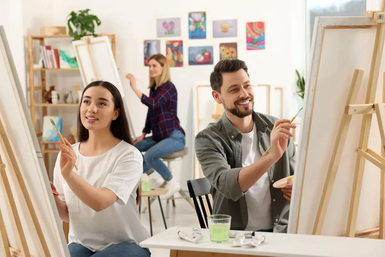 A couple sits side-by-side painting canvases with a painting lady behind.