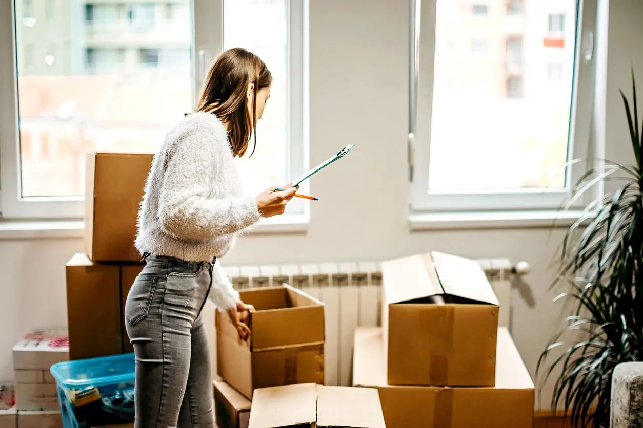 A young woman holding a clipboard examines items in cardboard boxes as she prepares to move.
