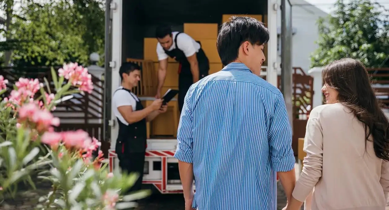 A smiling couple holds hands as professional movers check all the boxes in the truck.