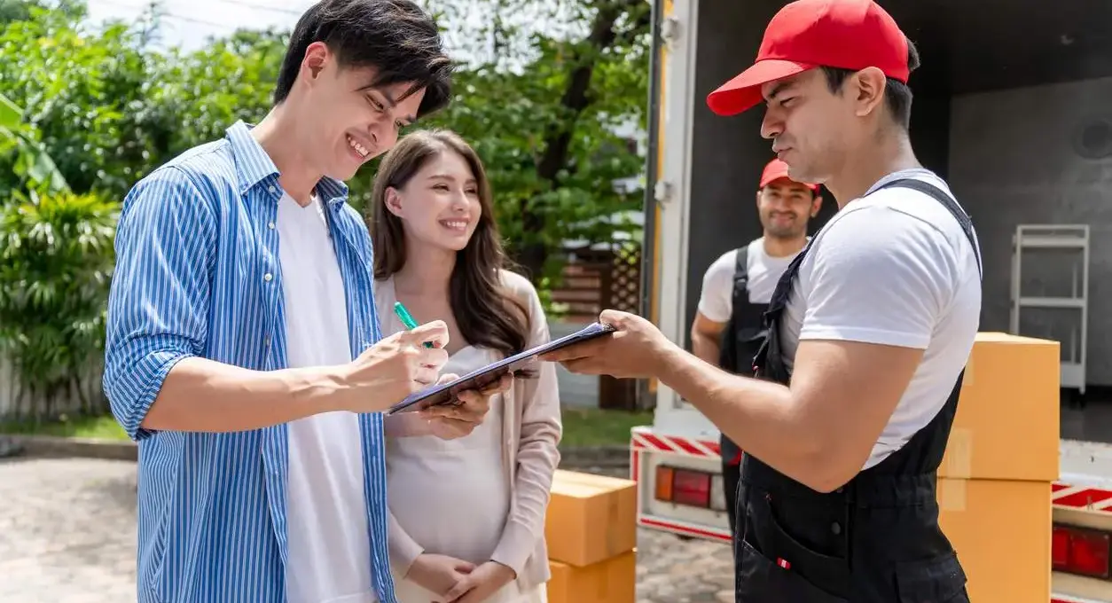 A smiling couple signs a clipboard from a duo of movers.