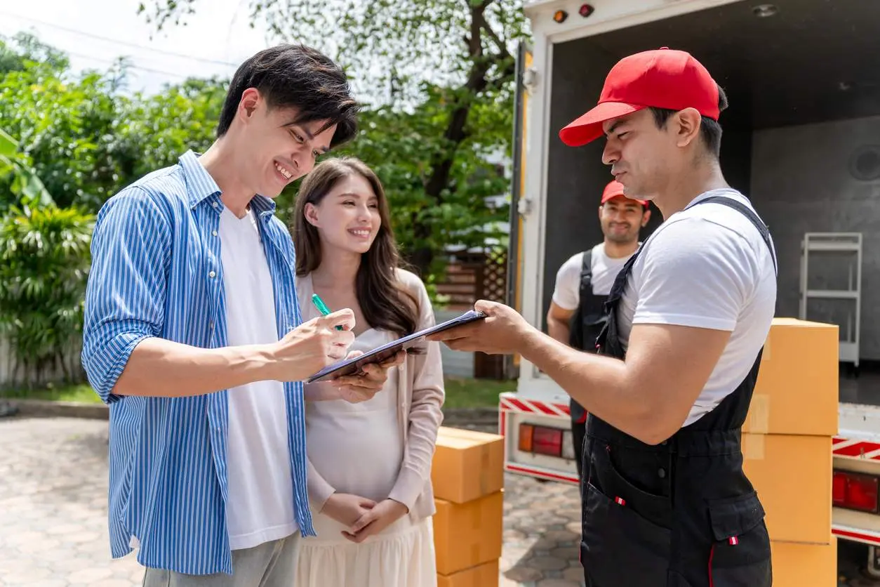 A smiling couple signs a clipboard from a duo of movers.