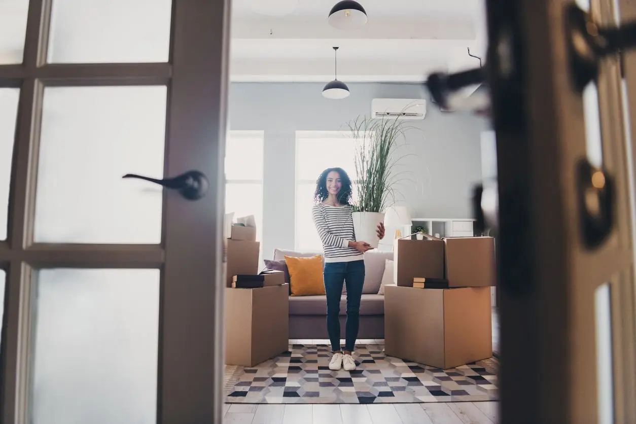 A smiling young woman stands in her new apartment surrounded by moving boxes and holding a plant.