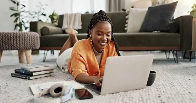 A smiling young woman lies on the floor typing on a silver laptop in a modern living room.