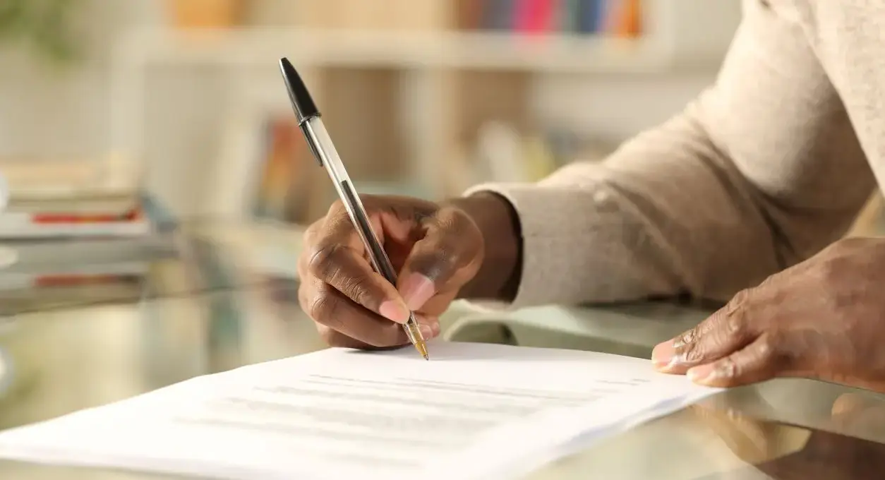 : A man signs a document on a reflective table with a black pen in his right hand.