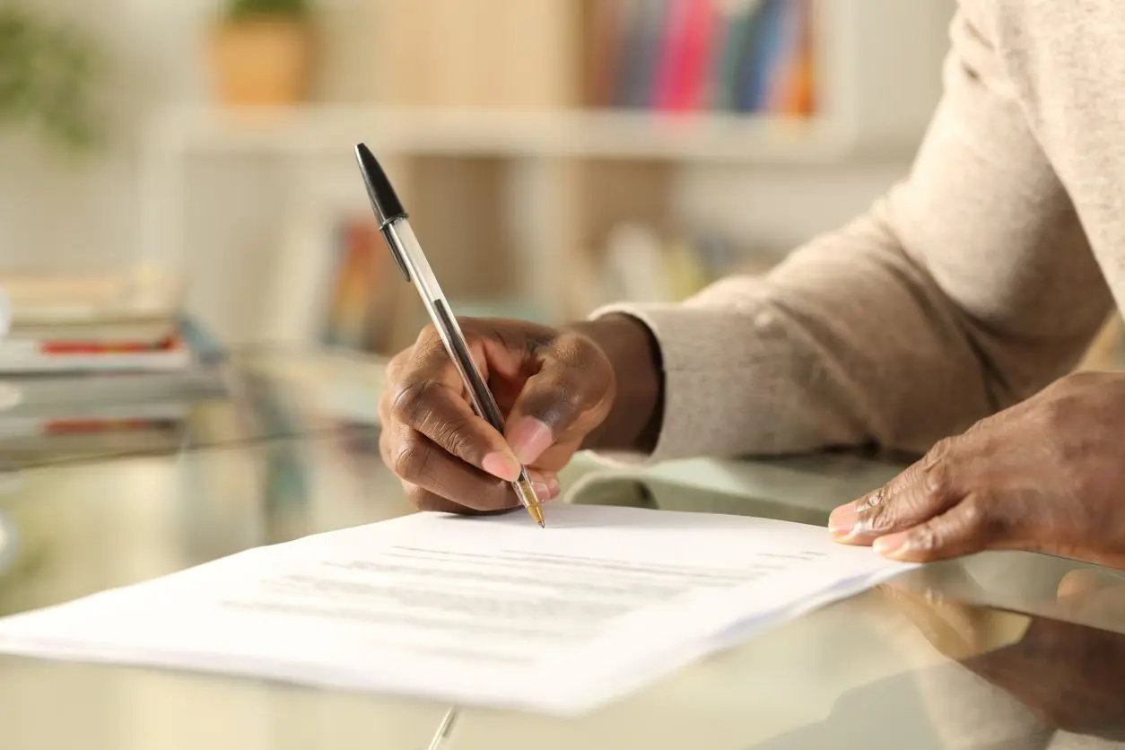 : A man signs a document on a reflective table with a black pen in his right hand.