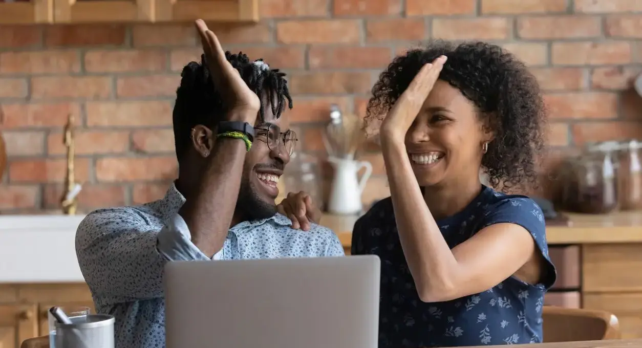 A happy, smiling couple high-five while sitting at a wooden table with a laptop open in front of them.