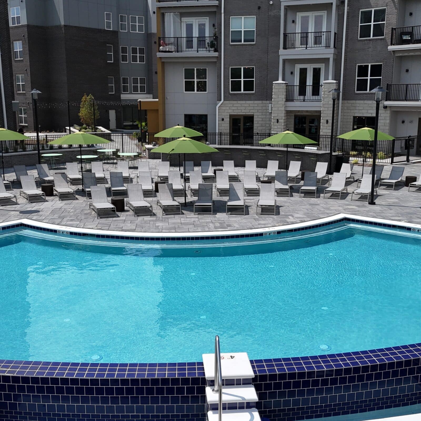 Modern apartment complex pool deck with a sparkling swimming pool, lounge chairs, and green umbrellas.