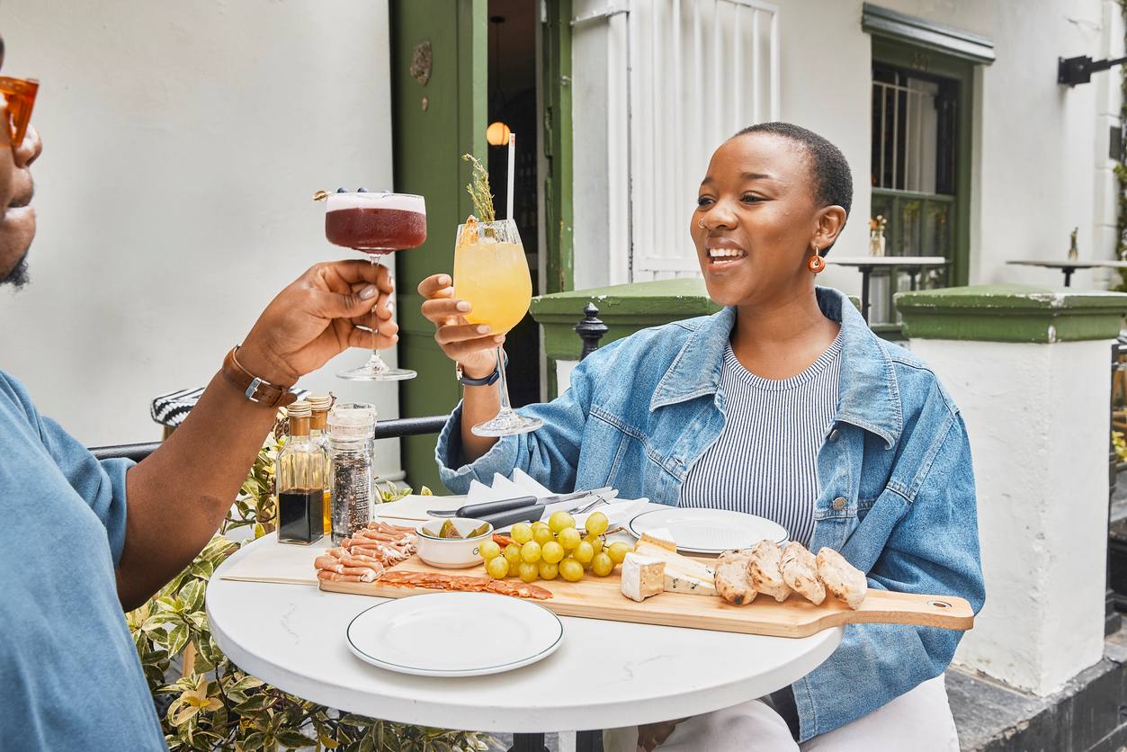 : A happy couple raises their cocktails in a toast during lunch outdoors, with a charcuterie board in front of them