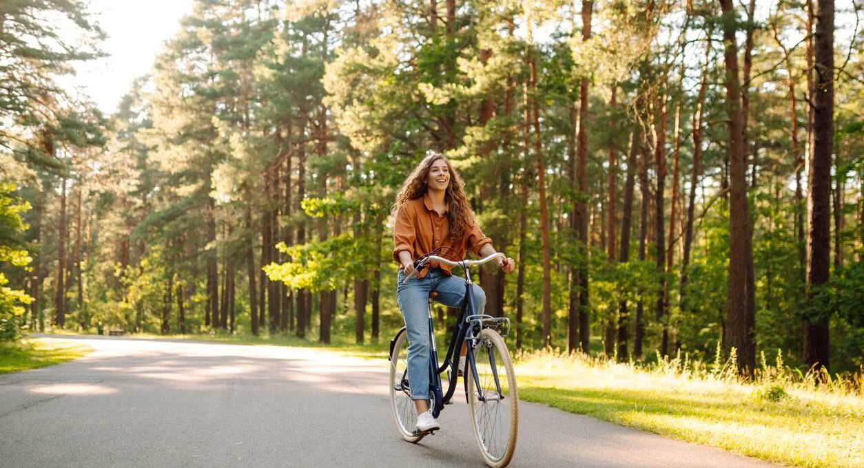 A smiling young woman rides a bike in the middle of a trail surrounded by trees on a sunny day.