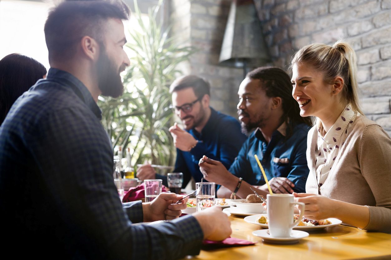 A group of happy friends eat and chat together, set against a painted brick wall and a large leafy plant.