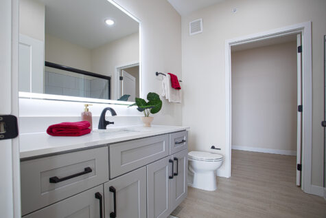 Modern bathroom at Relay 129 with sliding glass shower door, white quartz vanity, LED backlit mirror, matte black plumbing fixtures, and contemporary cabinetry.