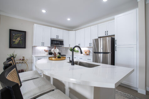 Modern kitchen at Relay 129 with white quartz island, stainless steel appliances, tile backsplash, large capacity sink, matte black plumbing fixtures, grey and white cabinetry, and island bar seating.