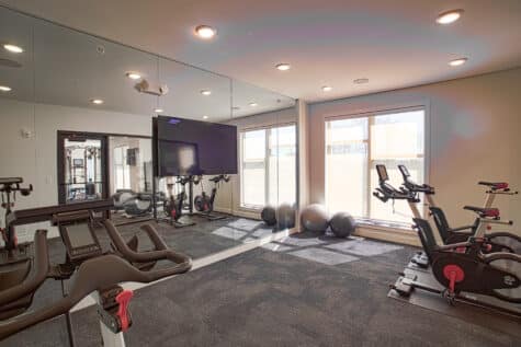 Fitness studio at Allure Apartments featuring stationary bikes, mirrored walls, mounted TV, and floor-to-ceiling windows with natural ligh