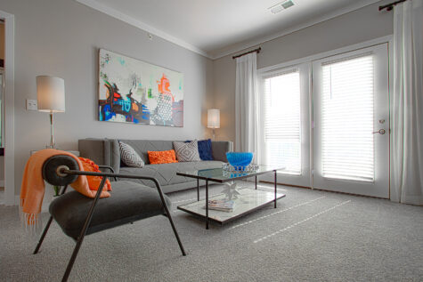 Contemporary living room at Allure in Centerville, Ohio by Hills Properties featuring plush carpeting, modern seating, colorful accent décor, large windows with natural light, and a private patio entrance.
