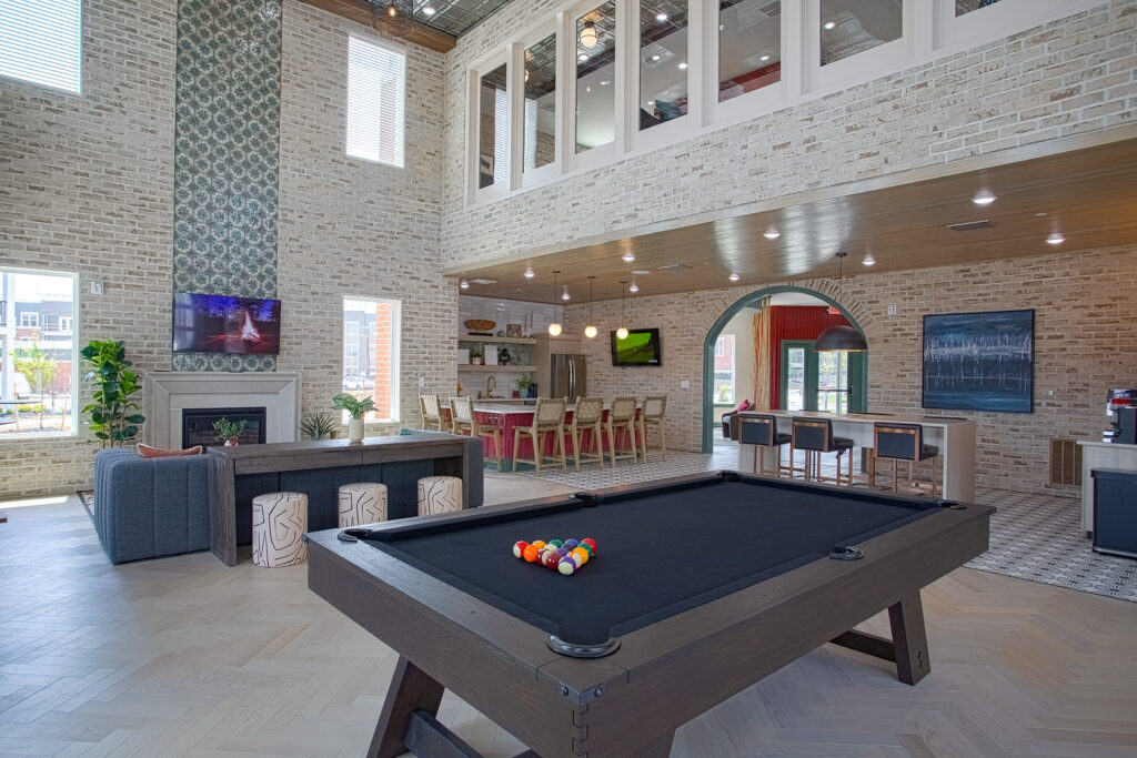 Spacious clubhouse lounge with billiards table, fireplace, open kitchen bar seating, and double-height brick walls with modern industrial décor.