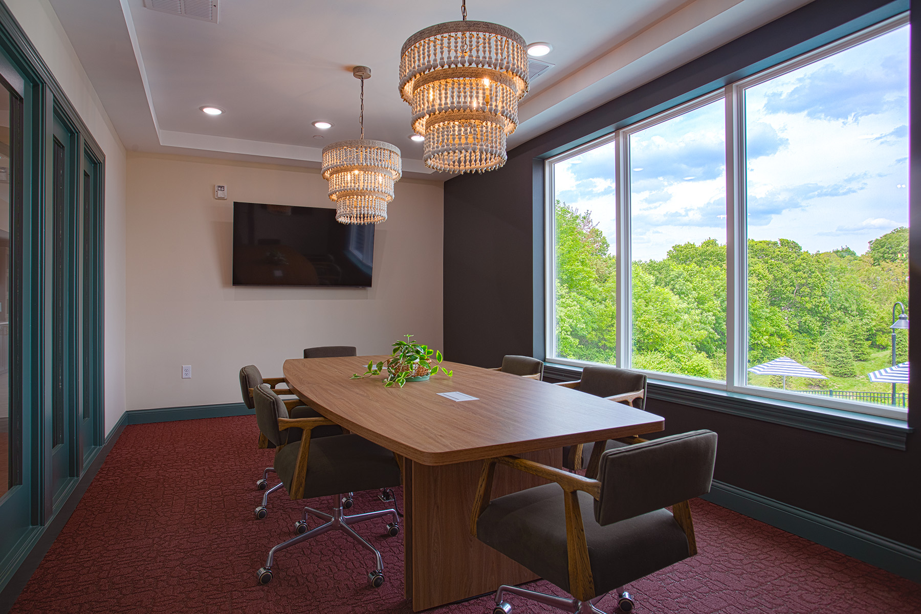 Contemporary conference room with wooden meeting table, upholstered rolling chairs, crystal chandeliers, and large windows overlooking trees.