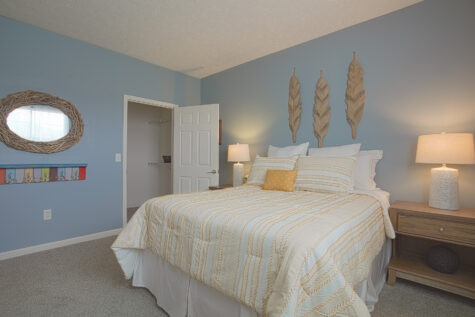 Spacious bedroom at Sterling Lakes Apartments in Mason, Ohio, with soft blue walls, plush carpeting, a queen-sized bed with neutral bedding, matching nightstands and lamps, decorative feather wall art, and an adjacent walk-in closet.