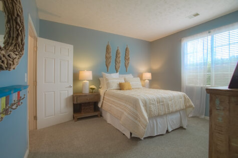 Bright primary bedroom at Sterling Lakes Apartments in Mason, Ohio, showcasing a large window with sheer curtains, plush carpeting, a queen-sized bed with textured neutral bedding, coordinating bedside tables and lamps, and calming blue accent walls.
