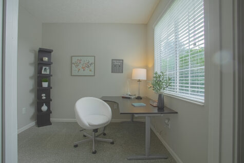 Dedicated home office at Sterling Lakes Apartments in Mason, Ohio, featuring a large window with blinds, plush carpeting, a modern desk with task lamp, a white swivel chair, neutral gray walls, and decorative shelving for a comfortable work-from-home setup.