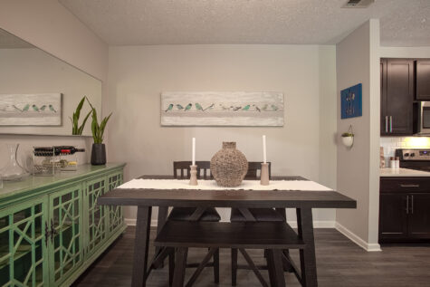 Modern dining area at Sterling Lakes by HILLS Properties featuring a dark wood table with bench seating, decorative centerpiece, green accent sideboard, wall mirror, and contemporary artwork, with an open connection to the kitchen.
