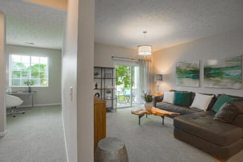 Bright open-concept living space at Sterling Lakes by HILLS Properties showcasing a sectional sofa, large abstract wall art, sliding glass door to a private patio, and an adjacent workspace with natural light from oversized windows.