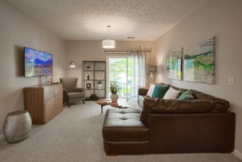 Inviting living room at Sterling Lakes by HILLS Properties showcasing a large sectional sofa, contemporary furnishings, wall-mounted TV, decorative shelving, and a sliding glass door leading to a private patio with green views.