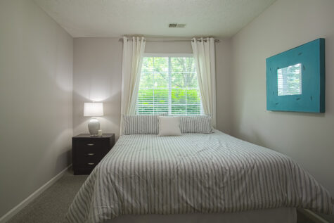 Bedroom at Sterling Lakes by HILLS Properties in Mason, Ohio featuring a bed with striped bedding, a nightstand with lamp, and a large window with views of mature landscaping.
