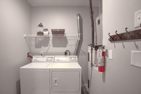 Convenient in-home laundry room at Sterling Lakes by HILLS Properties featuring a full-size washer and dryer, overhead storage shelving, utility hookups, and additional space for household organization and storage.