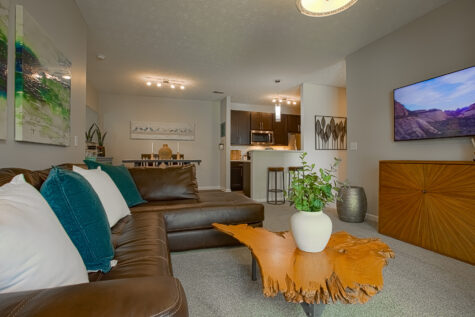 Open-concept living space at Sterling Lakes by HILLS Properties featuring a sectional sofa, natural wood coffee table, breakfast bar seating, modern kitchen with dark cabinetry, and a connected dining area ideal for entertaining.