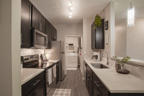 Modern galley-style kitchen at Sterling Lakes by HILLS Properties featuring dark espresso cabinetry, stainless steel appliances, quartz countertops, subway tile backsplash, and direct access to a spacious in-home laundry area.