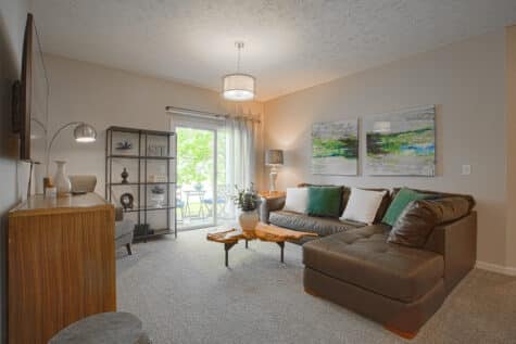 Bright and comfortable living area at Sterling Lakes by HILLS Properties featuring plush carpeting, a spacious sectional, modern artwork, accent lighting, and abundant natural light from the patio doors.