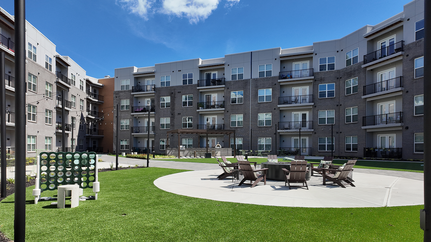 Landscaped resident courtyard at Vantage Anderson by HILLS Properties in Anderson, Ohio, featuring a community fire pit with Adirondack seating, oversized Connect Four game, outdoor grilling pavilion, and spacious green space surrounded by modern apartment buildings.