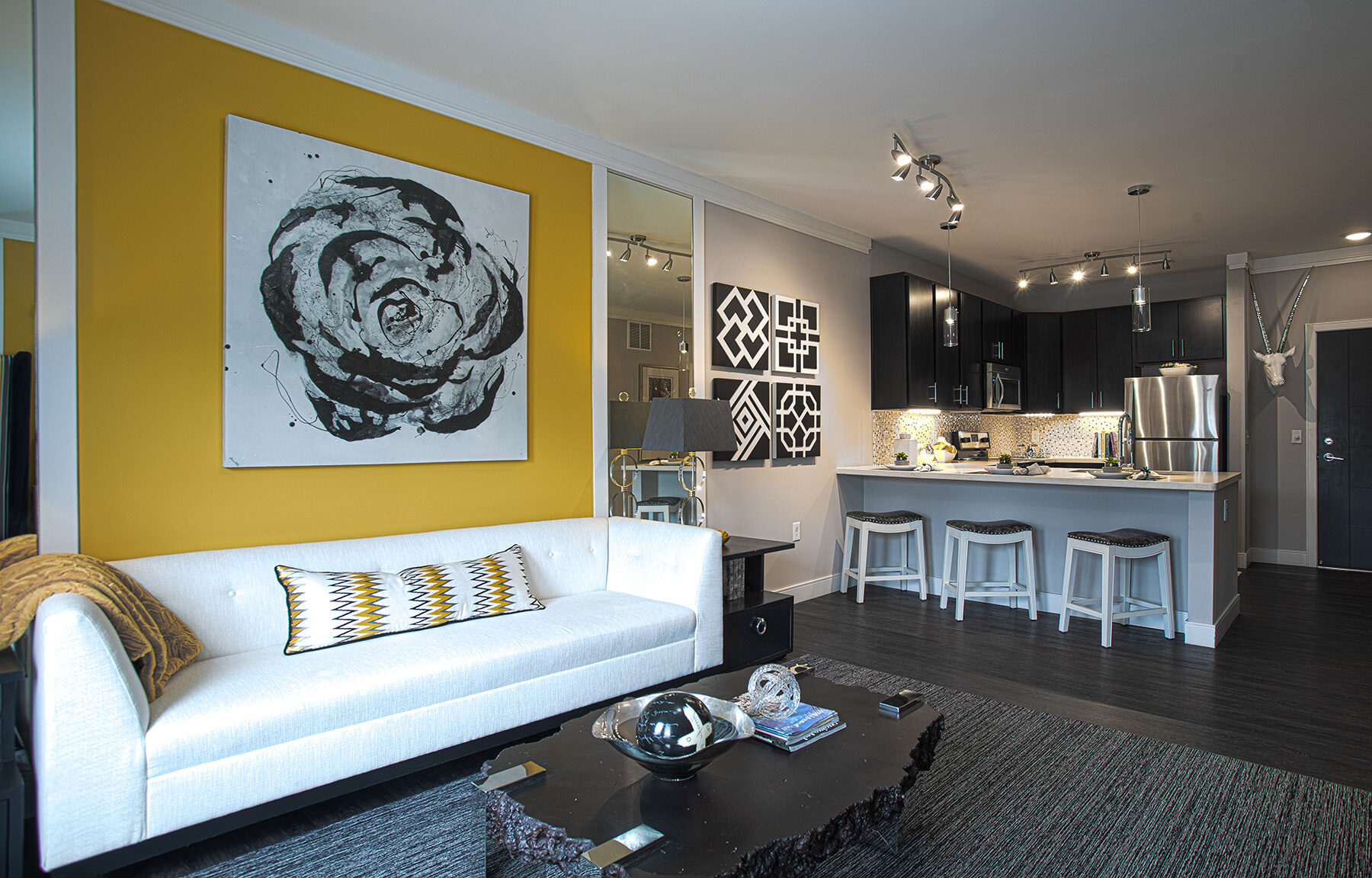 Open-concept apartment interior at Sterling Lakes in Mason, Ohio featuring a modern kitchen with dark cabinetry, stainless steel appliances, breakfast bar seating, pendant lighting, and an adjacent living room with contemporary furnishings and decorative artwork.